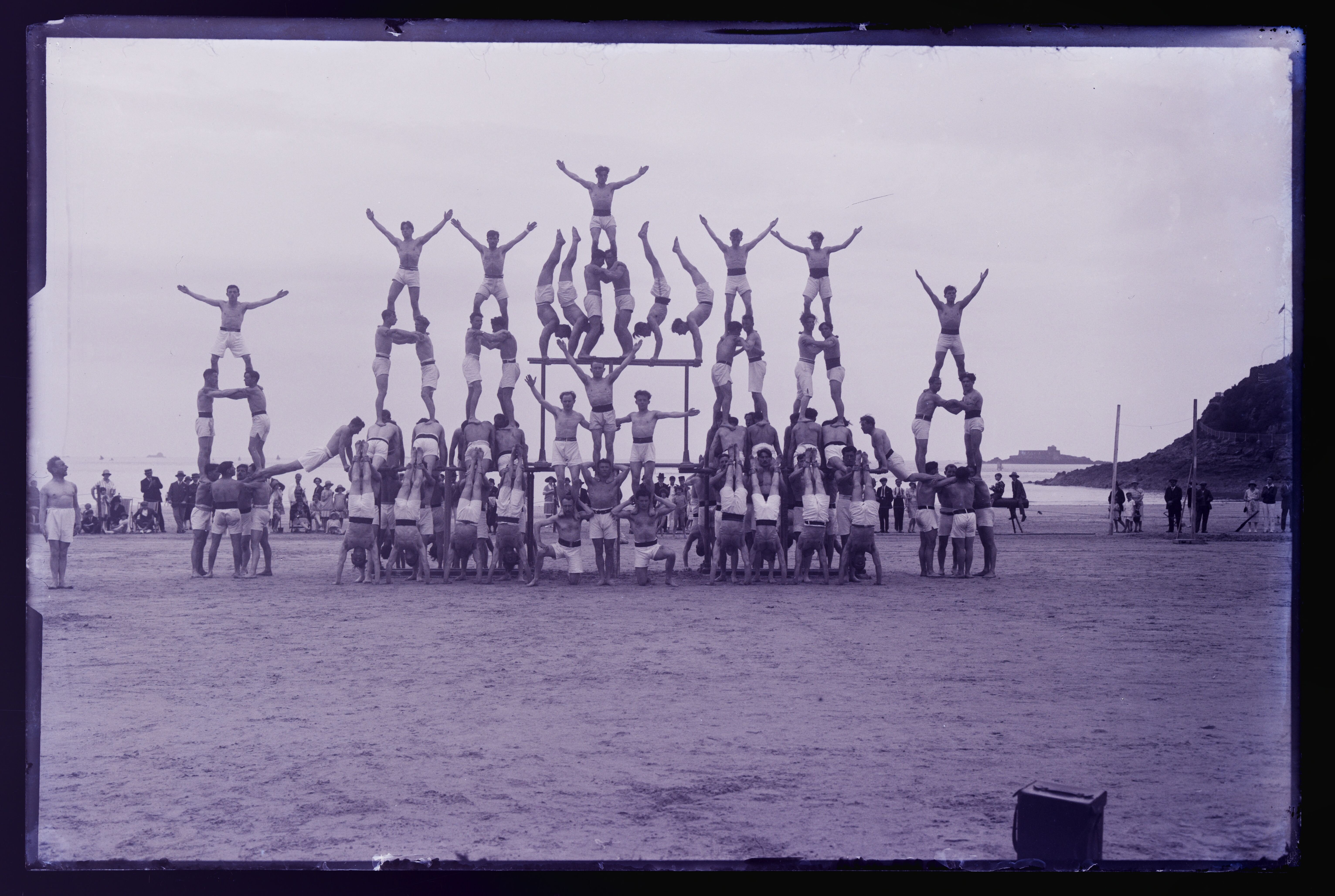 Rows of gymnasts on a beach forming several human pyramids of increasing height, watched by a crowd, with the sea and a small fort behind them.